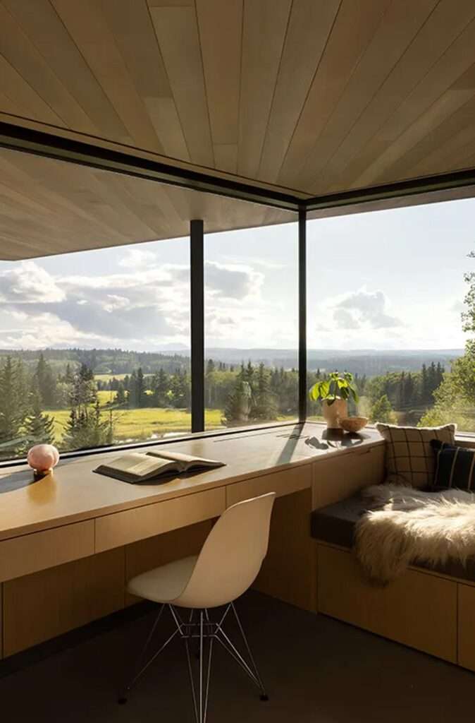 Minimalist home office desk integrated beside a cozy reading nook with sheepskin rug, looking out at an expansive evergreen valley.