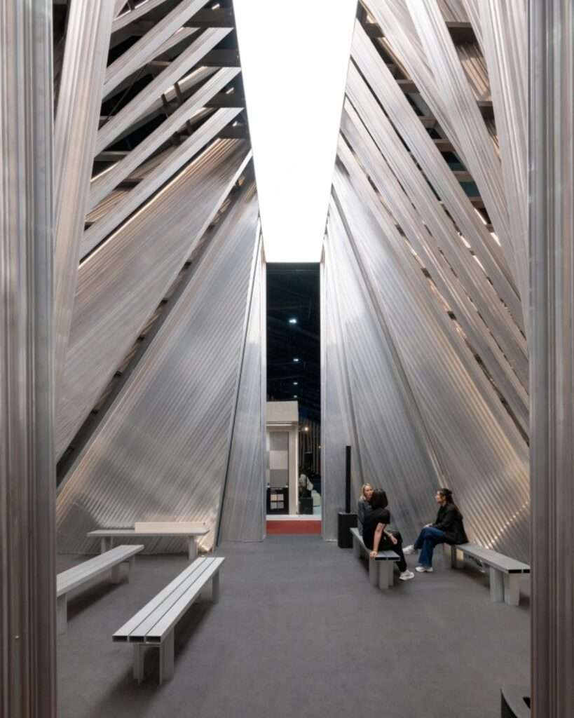 Axial interior shot looking down the central path of the UNFOLD pavilion with simple benches, metallic wall layers, and a vertical exit frame.