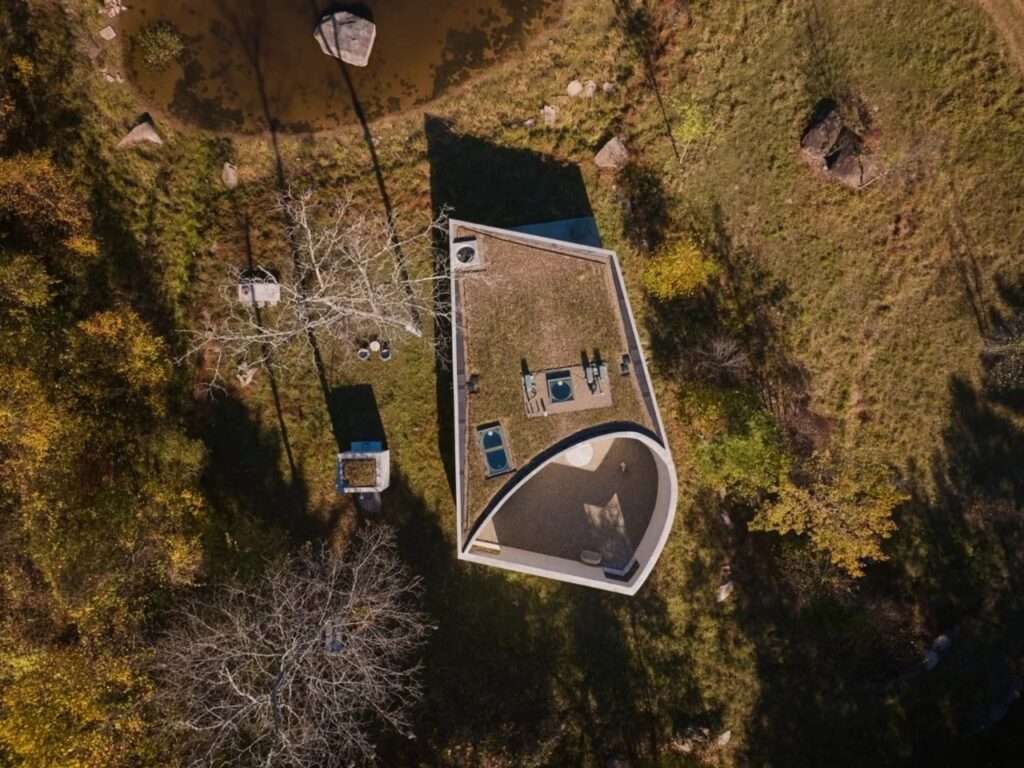 Aerial drone view of a geometric pavilion showing its green roof and curved courtyard embedded in a forest landscape.