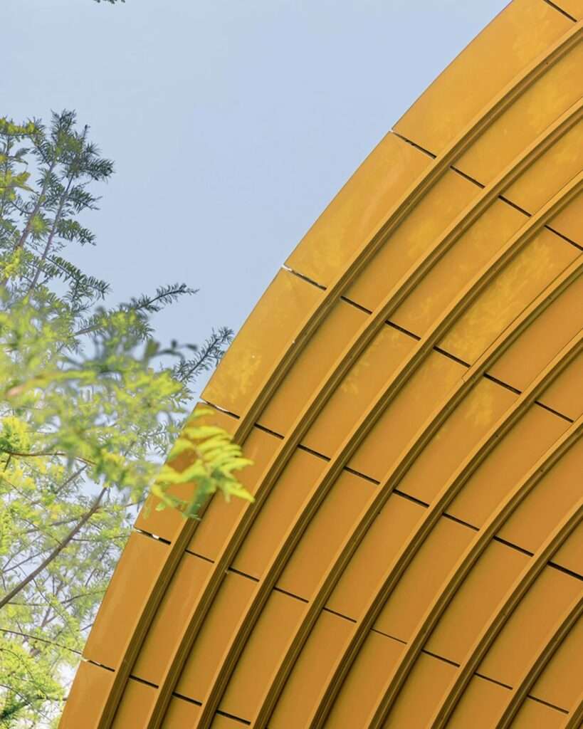 Elevated rear view of the Whale Gate pavilion showing the split blue shell, the golden staircase, and a person standing on the upper deck under a circular canopy.
