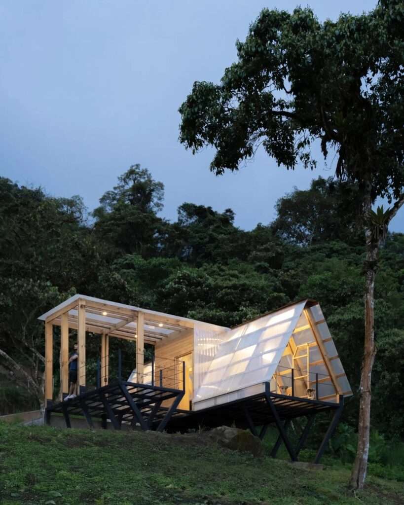 A side profile view of Casa 6-3's sharp triangular A-frame facade nestled behind an organic tree branch on a dirt hill overlooking distant green mountains.