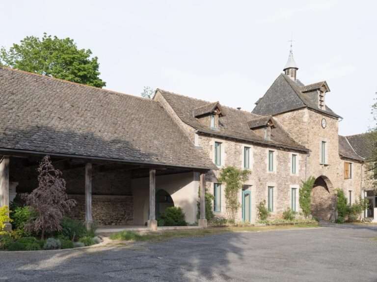 Exterior view of a renovated rustic stone farmhouse with an open timber porch, lush green courtyard planting, and a modern circular opening cut into the interior plastered wall.