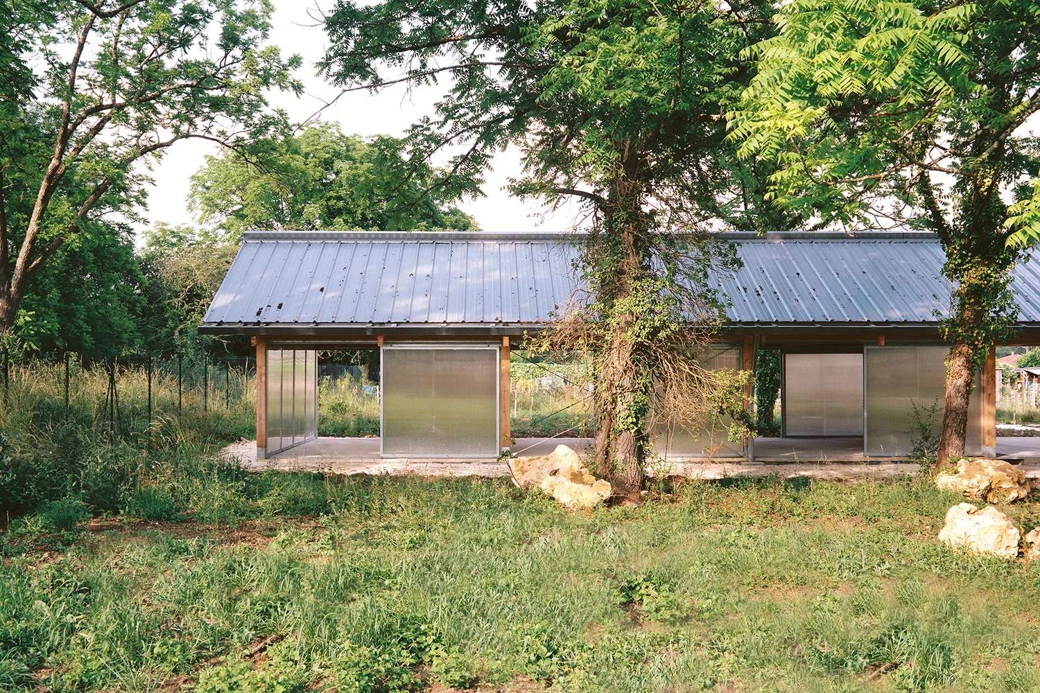 Longitudinal side profile view of the Gardeners' Shelter behind trees, showing the continuous translucent polycarbonate façade.