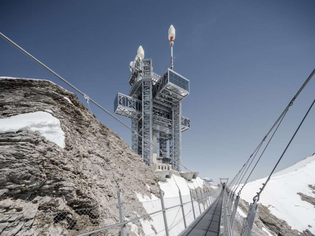 Metal suspension bridge leading towards the steel superstructure of the Titlis tower along a rocky mountain ridge.