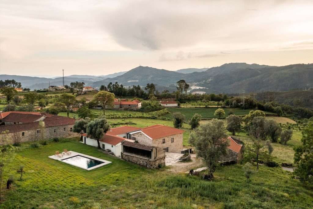 Aerial view of Abadim House showing its U-shaped layout, traditional terracotta tiled roofs, stone ruins, modern plaster volume, and an outdoor swimming pool set within a lush green mountainous landscape.