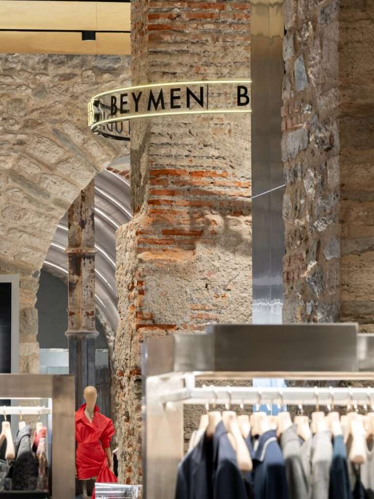 Close-up of a textured historic brick pillar with an illuminated circular Beymen sign attached, overlooking a modern clothing display.