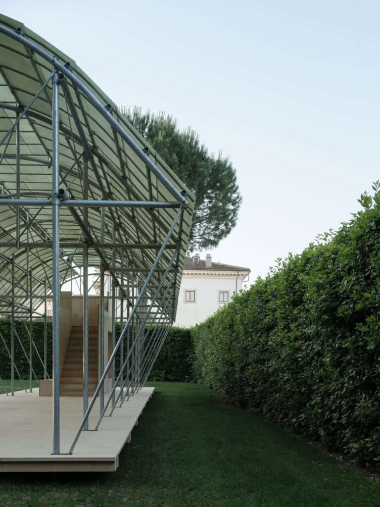 Side view of Cabane 7L architectural pavilion framing a view of the historic Villa Medici building behind high green hedges.