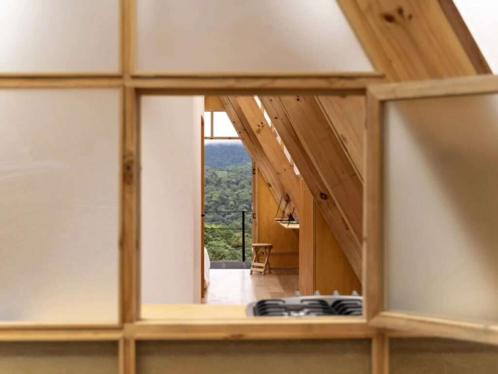 A detailed view looking out through an open square wooden window frame from the kitchen toward the interior hallway and distant green mountains.