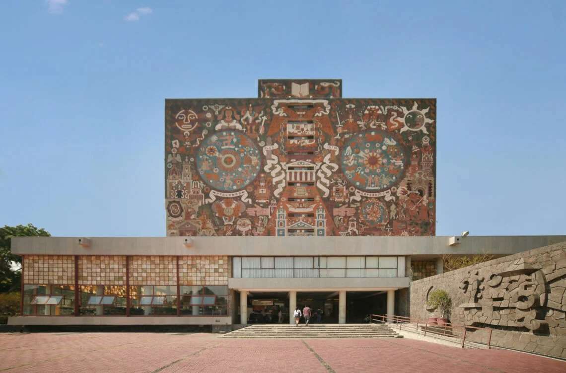 Dusk view of the UNAM Central Library showing the windowless upper tower illuminated from below against a dark blue evening sky, with the glass reading rooms glowing warmly from within.