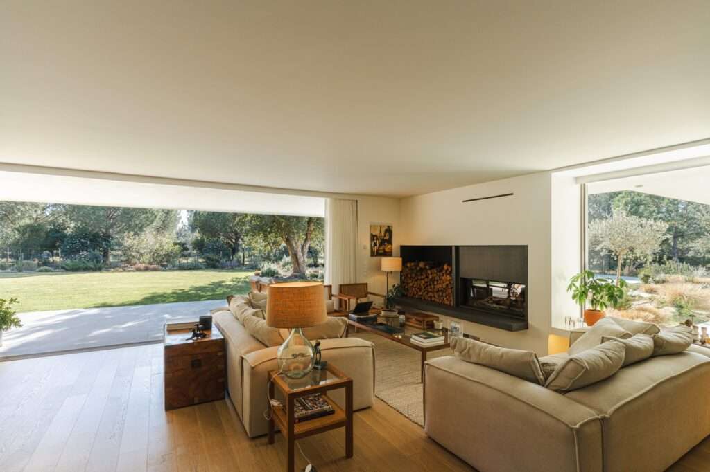 Panoramic view from inside the living room looking out onto a vast green lawn and surrounding forest through massive glass sliding doors.