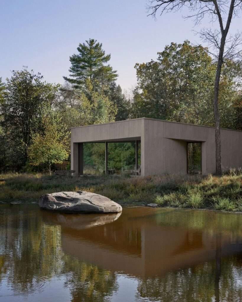 Angular concrete pavilion with glass facade reflecting in a pond containing a large prominent boulder.