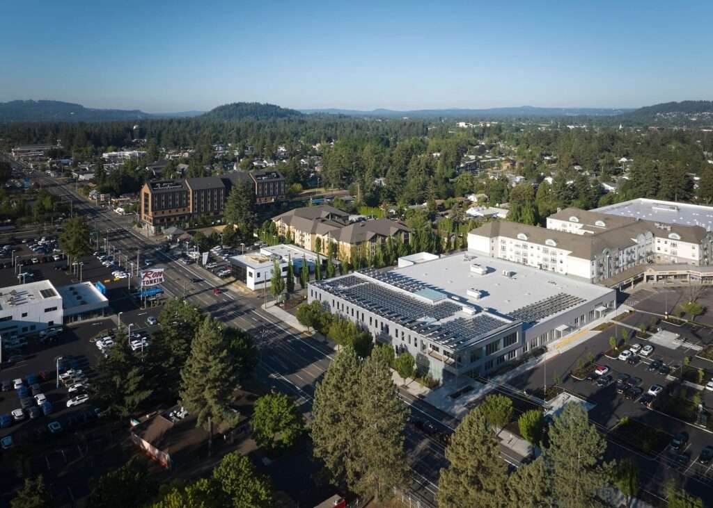 High-angle aerial photograph showing the expansive roof of the library operations center, covered with extensive solar arrays and skylights, positioned within a car-centric commercial suburban district in East Portland.