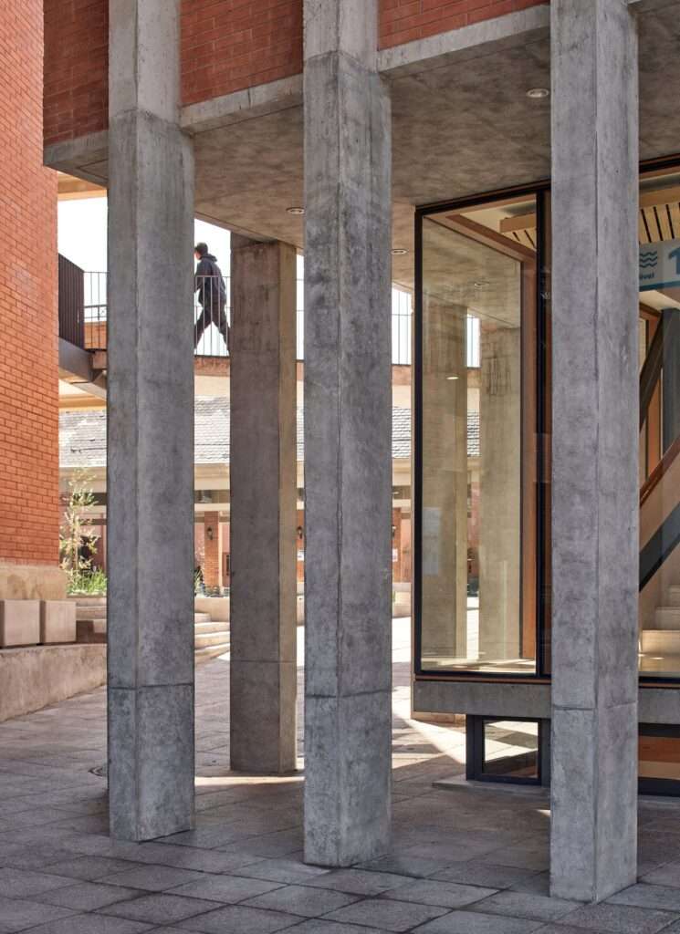 Close-up of raw concrete perimeter columns showing a pedestrian on an upper bridge and a recessed lower level utilizing the site's natural topography.