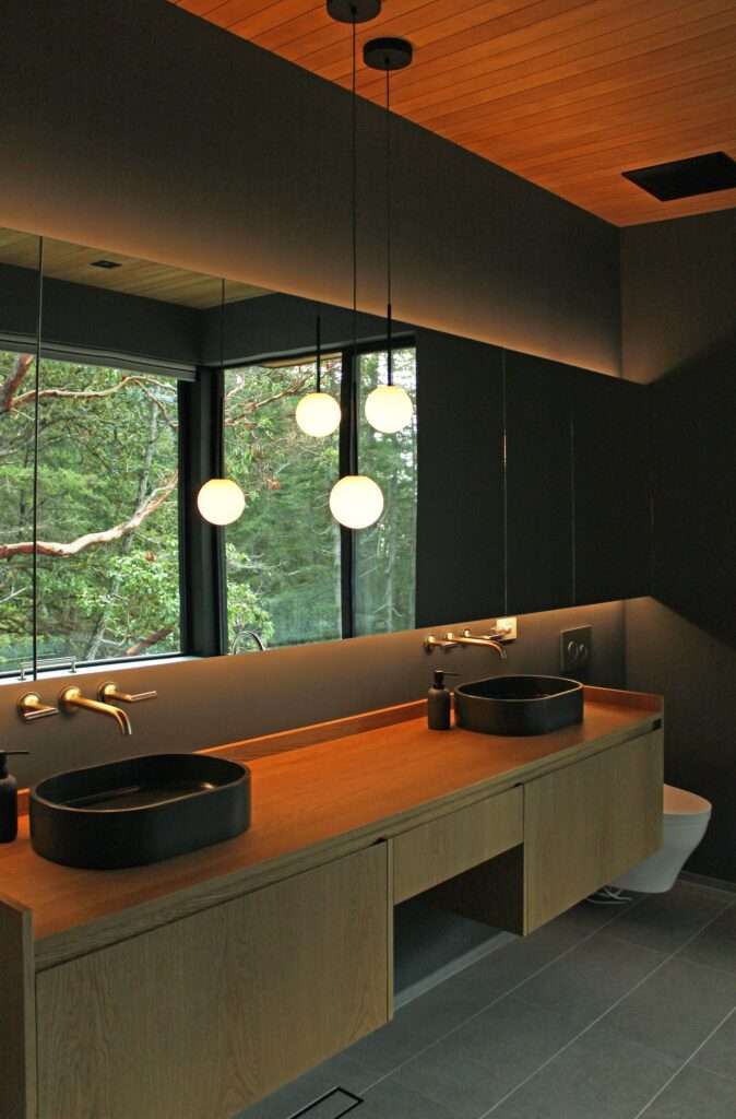 Luxury modern bathroom with long double wood vanity, black vessel sinks, and large mirror reflecting forest foliage window.