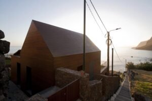 A low-angle exterior photograph of the Jardim do Mar Houses, showcasing the integration of the modern, gabled wooden structure on a steep coastal slope, held by massive, historic basalt stone terrace walls, with the Atlantic Ocean on the horizon under a clear sky.