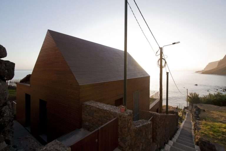 A low-angle exterior photograph of the Jardim do Mar Houses, showcasing the integration of the modern, gabled wooden structure on a steep coastal slope, held by massive, historic basalt stone terrace walls, with the Atlantic Ocean on the horizon under a clear sky.