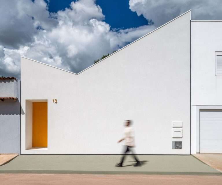 Minimalist stark white street front facade of a modern house under a dramatic blue cloudy sky, featuring an asymmetrical butterfly roofline and a recessed yellow entry door with a blurred person walking past.