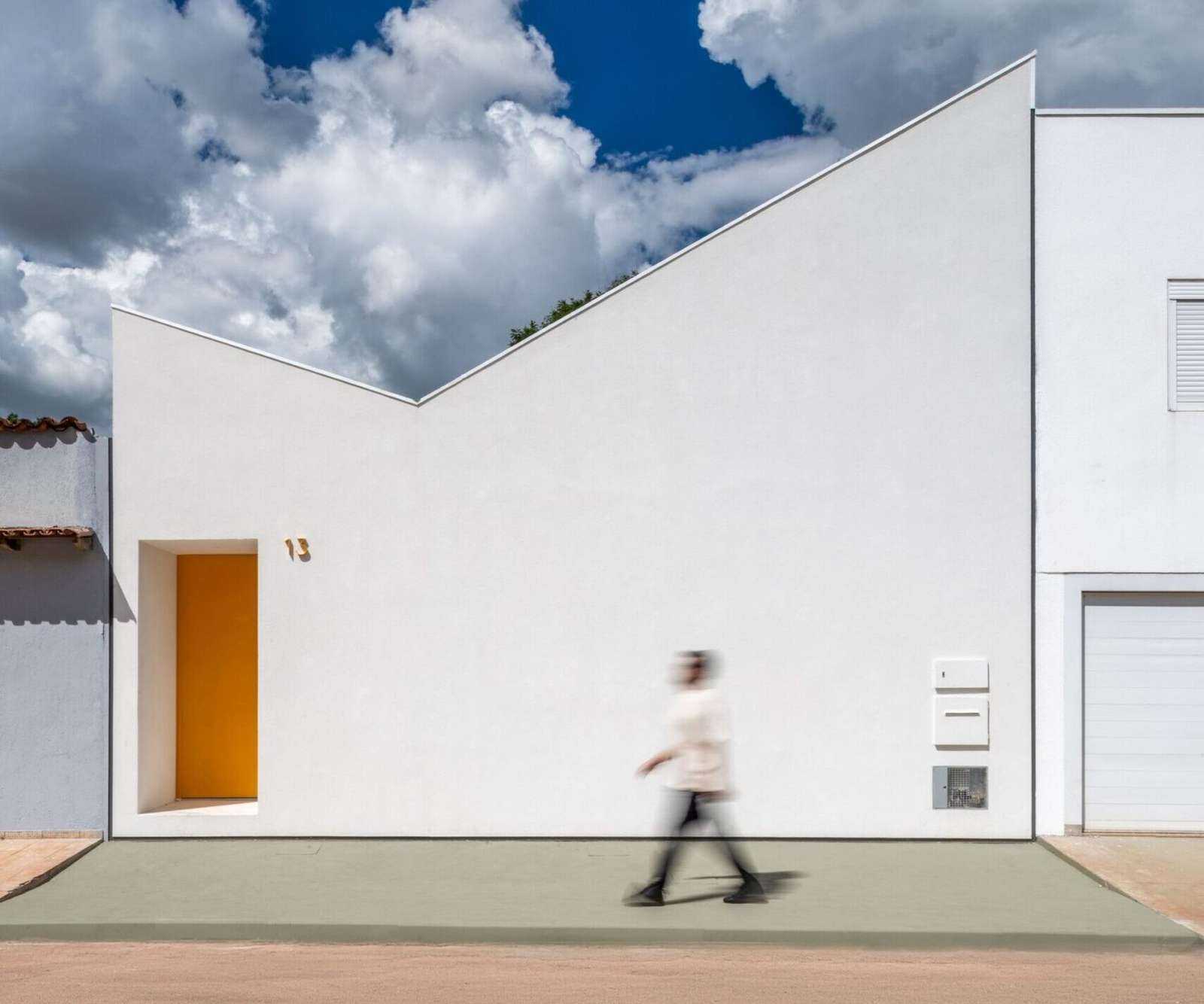 Minimalist stark white street front facade of a modern house under a dramatic blue cloudy sky, featuring an asymmetrical butterfly roofline and a recessed yellow entry door with a blurred person walking past.