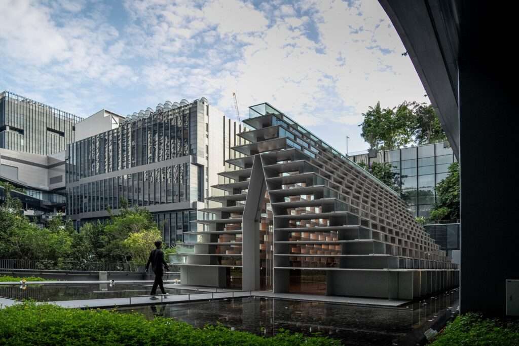 Exterior view of the glass meditation pavilion standing in a shallow water feature with a pedestrian walking nearby.