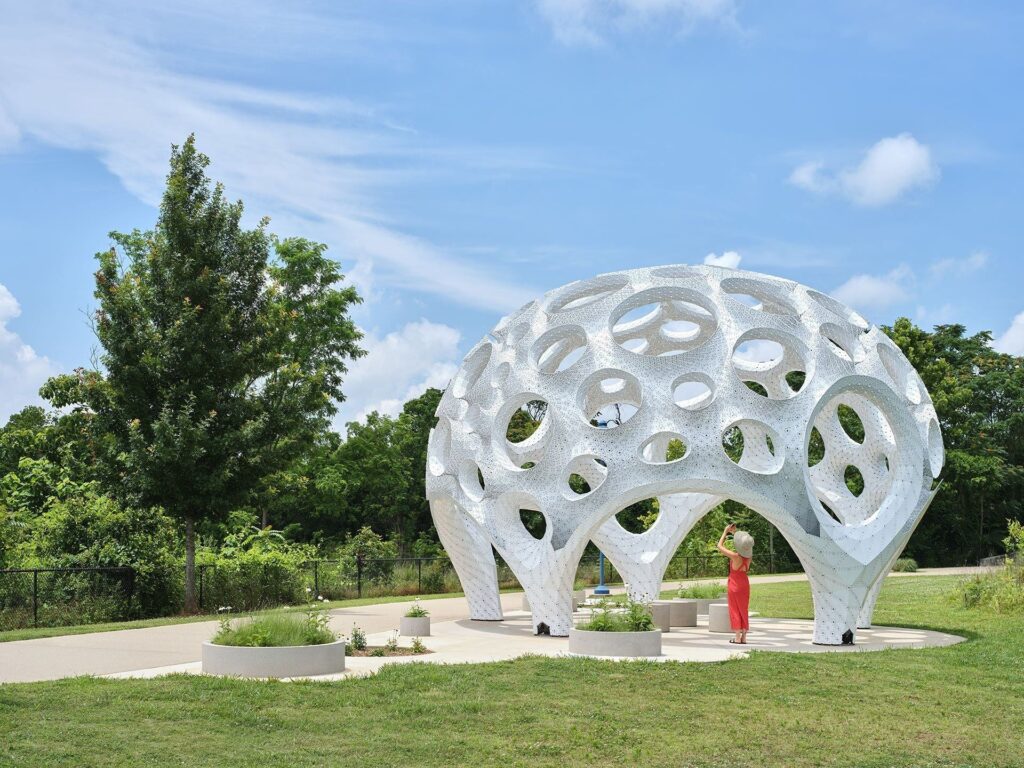 A side-angle shot of the white organic dome structure Moonrise by MARC FORNES / THE VERY MANY under a blue sky, featuring a woman in a red dress standing underneath taking a photo.