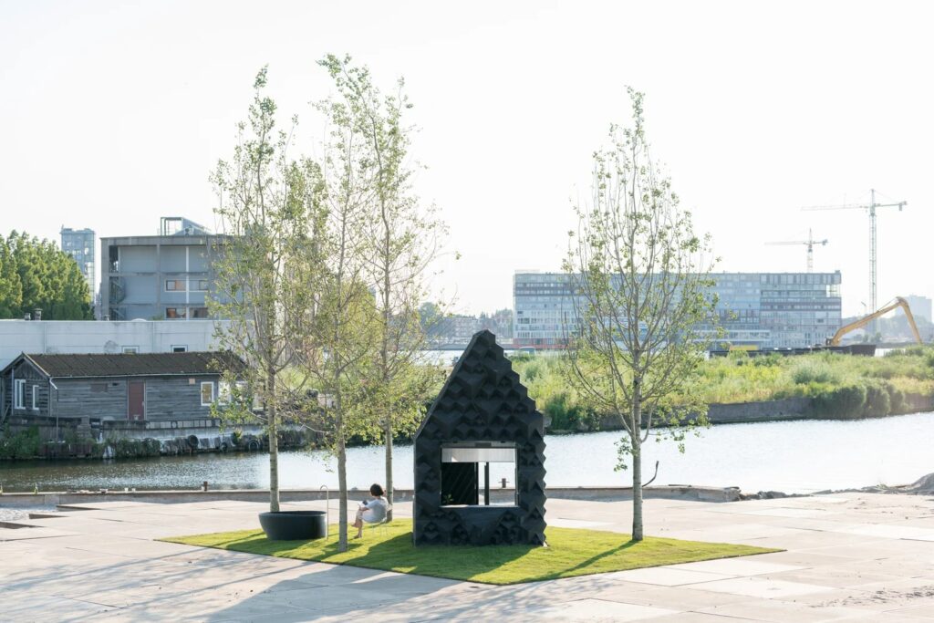 A small black 3D-printed micro-cabin on a patch of green grass next to a river canal in an industrial area of Amsterdam.