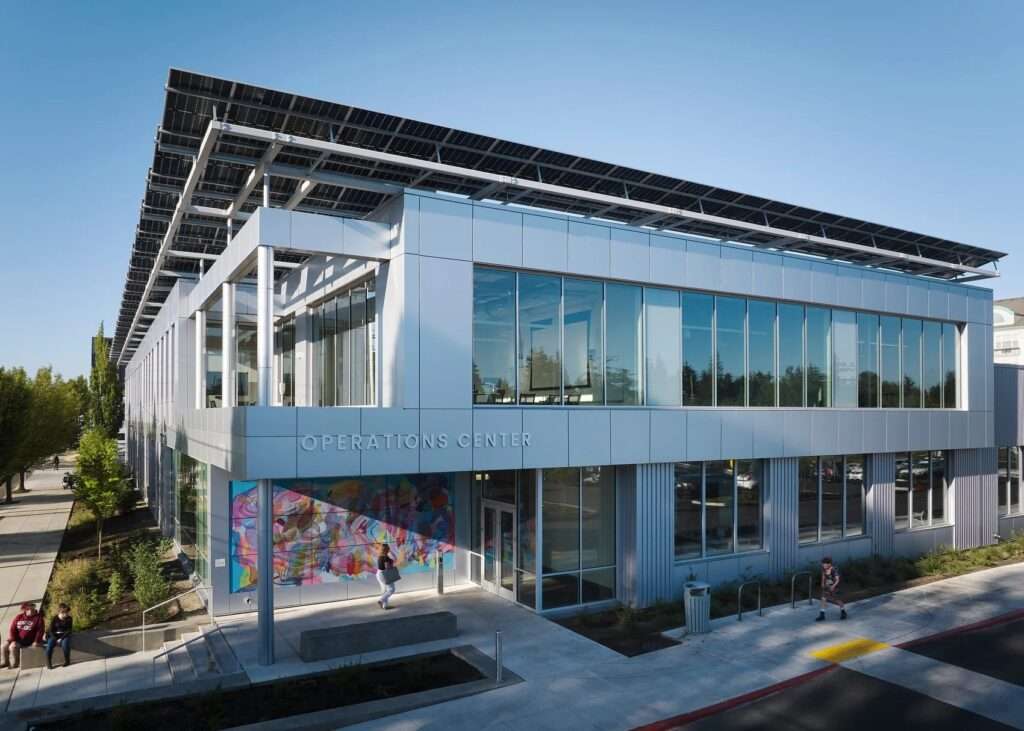Modern exterior of the Multnomah County Library Operations Center featuring a silver metallic facade, extensive glazing, a colorful vibrant mural at the ground floor entrance, and a large rooftop solar panel canopy under a clear blue sky.