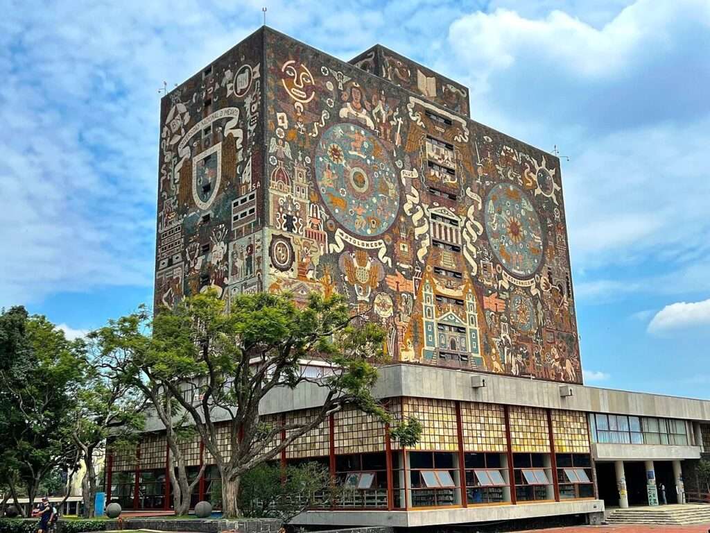 Symmetrical eye level front view of the main entrance to the UNAM Central Library with students walking up the steps beneath the massive mosaic archive tower and next to a carved volcanic stone relief wall on the right.