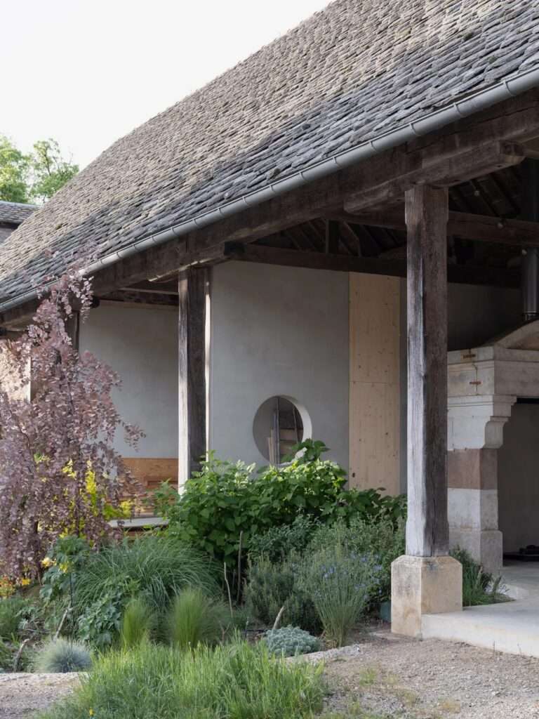 Dramatic double-height interior under original weathered timber roof trusses, featuring stepped wooden seating platforms, a suspended black metal fireplace, and a circular wall aperture.