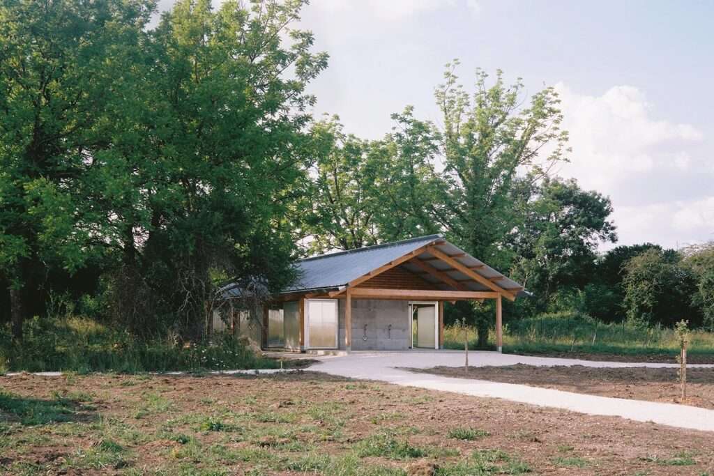 Angled exterior view of the Gardeners' Shelter showing the pitched metal roof, wooden columns, and polycarbonate sliding panels surrounded by lush trees.