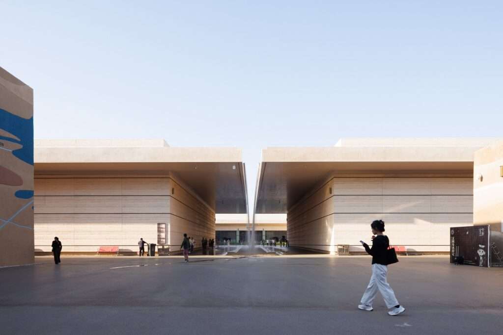 Daytime wide-angle shot of a deep architectural canyon walkway framed by monumental textured walls with visitors walking under a clear blue sky.