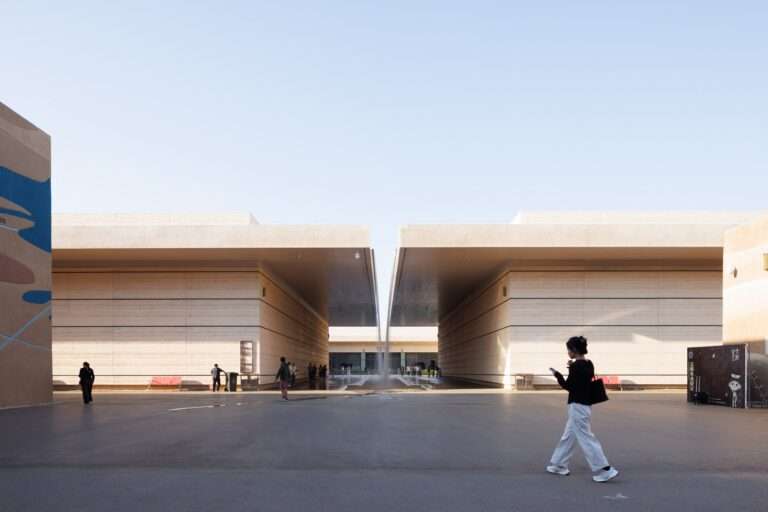 Daytime wide-angle shot of a deep architectural canyon walkway framed by monumental textured walls with visitors walking under a clear blue sky.