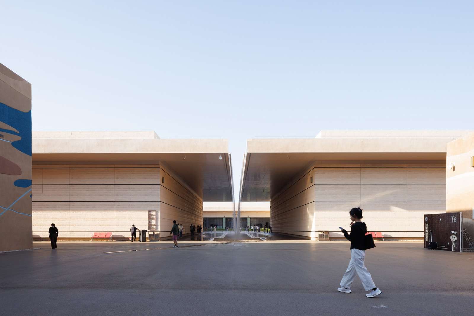 Daytime wide-angle shot of a deep architectural canyon walkway framed by monumental textured walls with visitors walking under a clear blue sky.