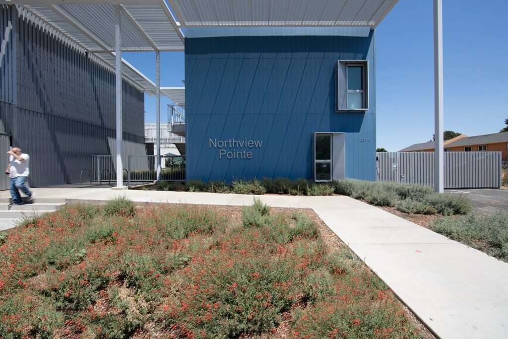 Ground-level view of a grassy courtyard at Northview Apartments with a black patio umbrella, outdoor seating, and colorful building facades under a clear blue sky.