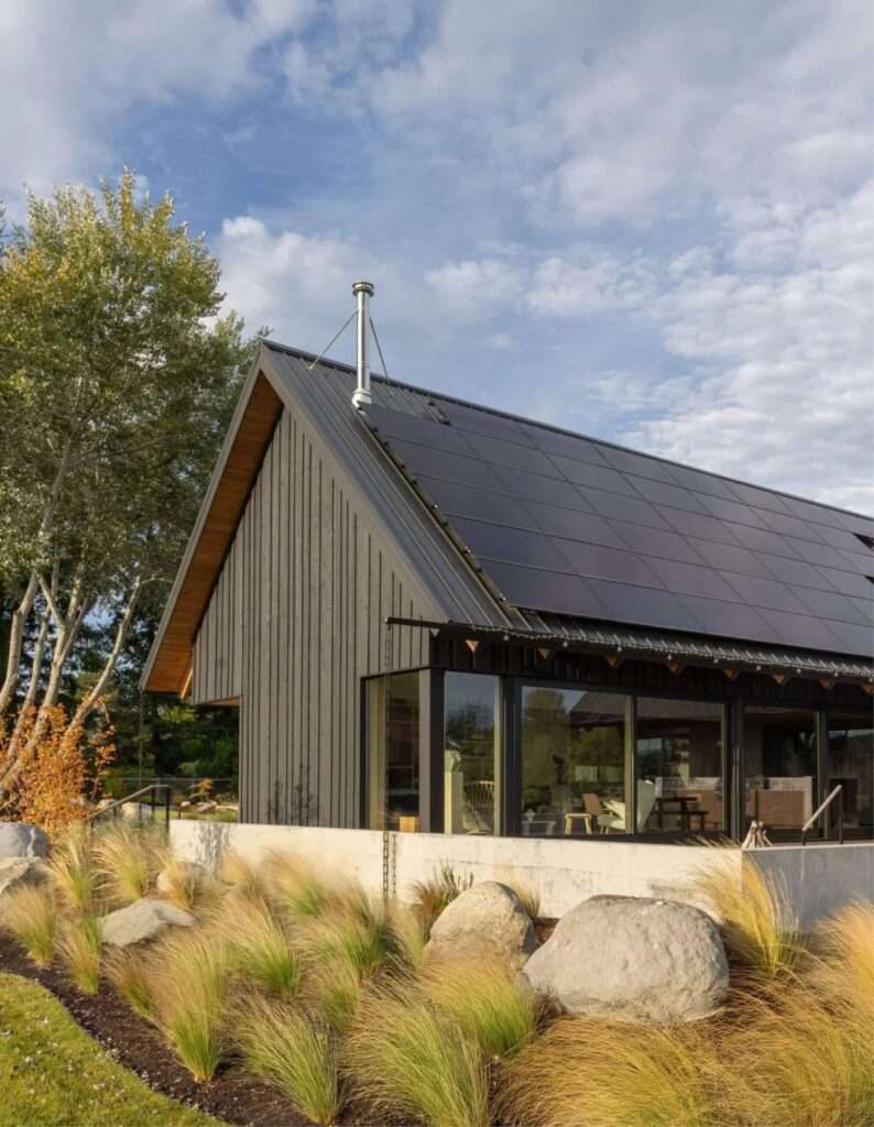 Full North facade of Day's Inlet House with dark vertical wood siding, landscaped native grasses, and stone pathways.