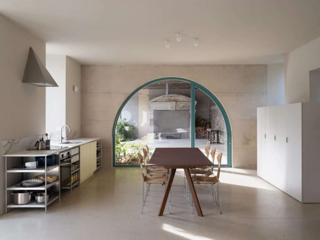 Minimalist farmhouse interior with light ash wood wall panels, exposed white ceiling beams, a large hollow circular wall opening, and a preserved antique green clockwork mechanism.
