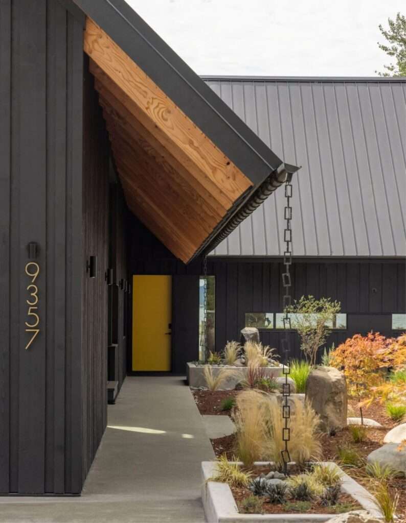 Covered entry walkway along the dark timber wall with exposed natural wood roof rafters and a yellow door.
