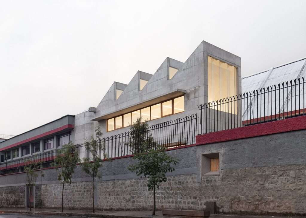 Street-side view of the sawtooth concrete arts building rising behind an old stone retaining wall topped with a black metal fence under an overcast sky.