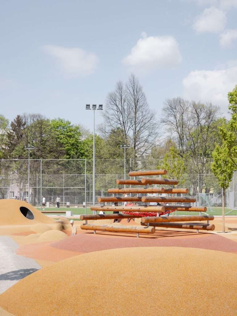 Wooden climbing frame in a school playground featuring a red net, set against a backdrop of trees and a fenced sports area.