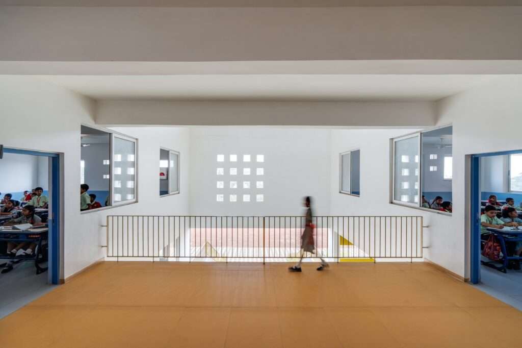First-person view of a school corridor overlooking an atrium with large windows opening into classrooms on both sides and a student walking past a metal railing.