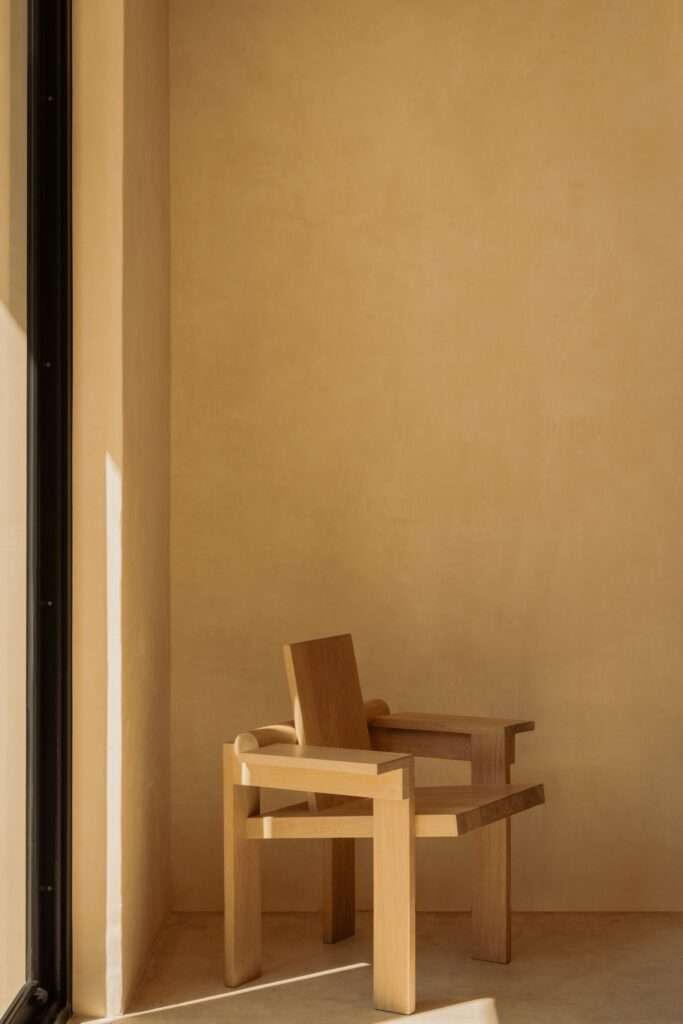Sculptural solid wood armchair positioned in a corner with sunlight streaming across the chukum plaster wall and concrete floor.