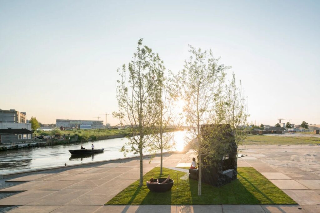 Wide sunset view of the 3D-printed Urban Cabin by a river with a person relaxing in an outdoor bathtub next to a passing boat.