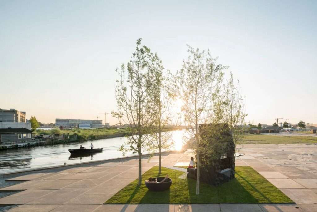 Wide sunset view of the 3D-printed Urban Cabin by a river with a person relaxing in an outdoor bathtub next to a passing boat.