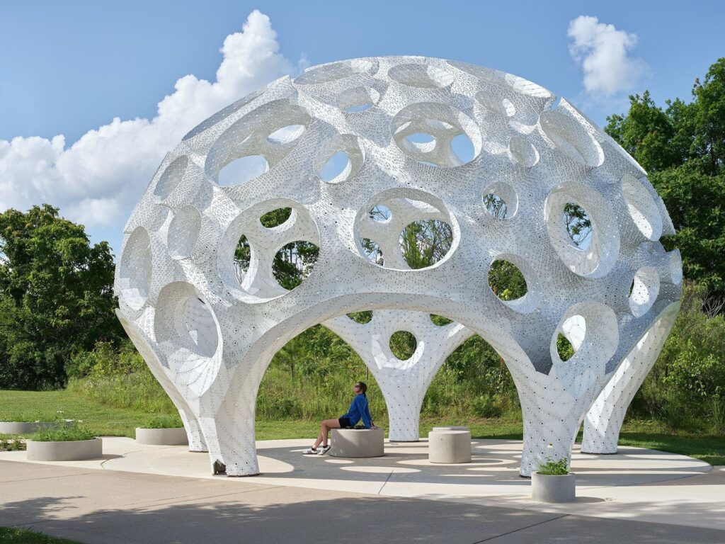 Eye-level view of the Moonrise ultra-thin aluminum perforated pavilion structure with a visitor sitting on a concrete bench inside looking up.