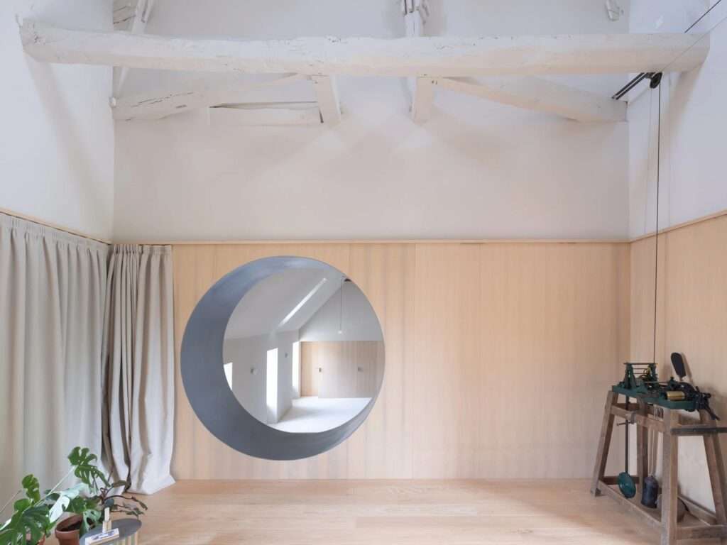 Close-up frame peering through a smooth, thick circular wall opening into a sunlit reading alcove with built-in wooden bookshelves and green potted plants.