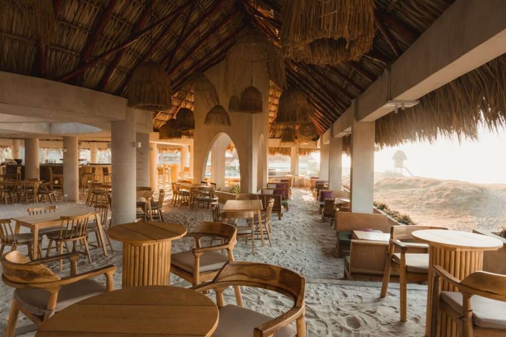 A wide view of an open-air beach club pavilion with a massive timber thatched roof, filled with wooden tables, woven pendant lights, and sandy floors leading toward a sunlit beach.