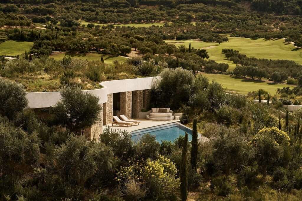 Aerial view of a subterranean stone villa with a green roof, infinity pool, and sun deck surrounded by olive groves and a golf course.