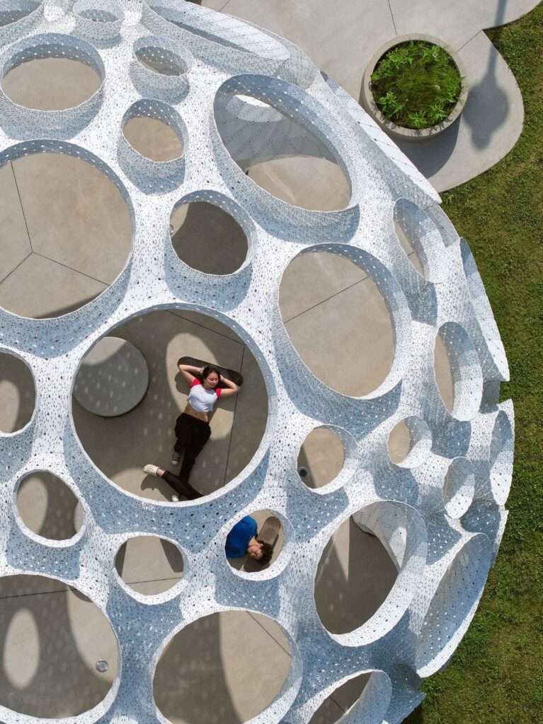 Top-down drone view looking into the perforated white pavilion dome, showing two individuals lying down inside with skateboards.