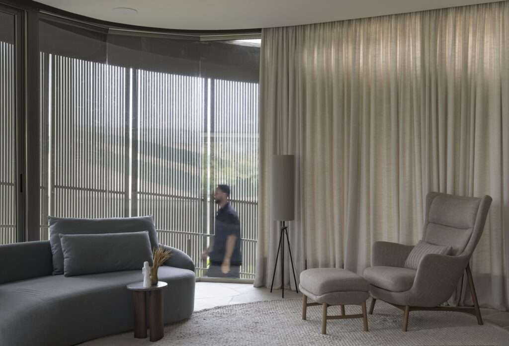 Close-up of an indoor lounge area at Casa Circular next to a curved glass window wall, showing a silhouette of a person walking along the external ramp behind vertical brise-soleil slats.