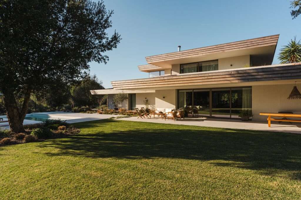 Exterior view of the two-story horizontal villa from the expansive green lawn, highlighting Capoto plaster finishing and natural wood pergolas.