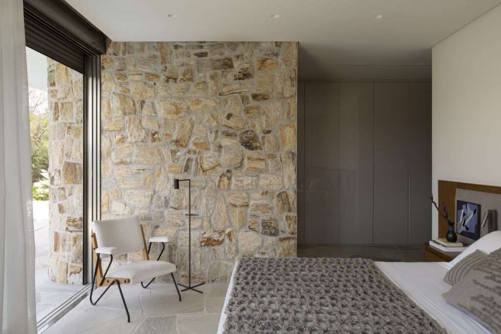 Minimalist master bedroom interior in Casa Circular with a prominent rustic stone accent wall, large floor-to-ceiling glass doors, a white accent chair, and a neat neutral bed setup.